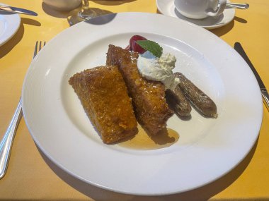 Krem şanti ve nane soslu çıtır Fransız tostu, sosis bağlantıları, şurup çiselemesi ve sarı masa örtüsü üzerine konmuş çilek sosu içeren bir tabakta..