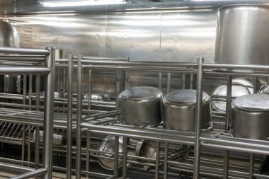 Geniş metal tencere ve tavaları tutan paslanmaz çelik rafları olan, parlak tepedeki aydınlatma altında yansıtıcı metal duvarlarla çevrili bir ticari mutfak deposu..