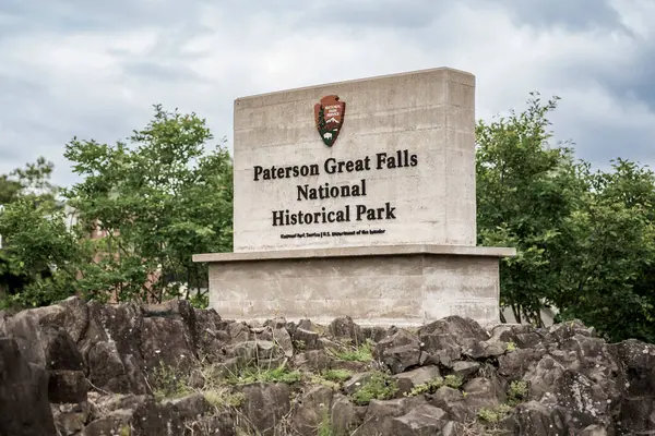 Paterson, NJ - ABD - 24 Mayıs 2025 Paterson Büyük Şelaleleri Ulusal Tarih Parkı tabelası bulutlu bir gökyüzü altında engebeli kayalar ve yeşillikler arasında duruyor..