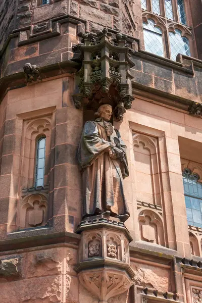 Princeton, NJ - ABD - 31 Mayıs 2025 Gotik Princeton binasında süslü bir tepe örtüsü, kemerli pencereler ve karmaşık mimari oymalar tarafından çerçevelenmiş önlüklü taştan bir figür duruyor..