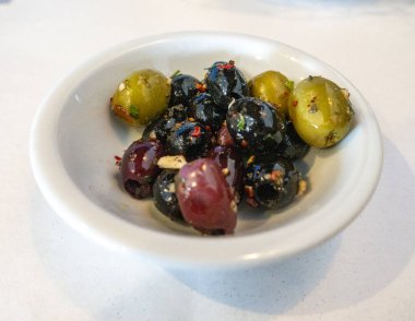 Küçük beyaz bir tabakta ezilmiş sarımsak ve kırmızı biber taneleriyle vurgulanmış yağ ve otlarla marine edilmiş yeşil, siyah ve mor renkli bir karışım bulunur. Lezzetli bir Akdeniz ısırığı