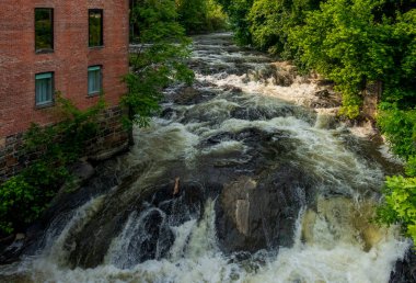 Beacon, New York - ABD 13 Haziran 2025 Beacon, New York 'taki ikonik Roundhouse Oteli' nin arkasında şiddetli şelaleler engebeli kayalara çarpıyor ve bu tarihi nehir kenarı inzivasına dramatik bir doğal zemin oluşturuyor..