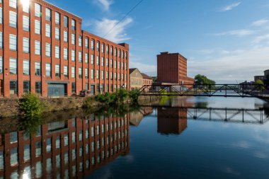 Holyoke, MA-ABD - 24 Ağustos 2025 Sakin bir kanal, kırmızı tuğlalı sanayi binalarını ve Holyoke 'de dağınık mavi bulutların altında bir köprüyü yansıtır.