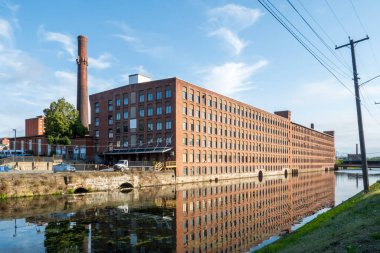 Holyoke, MA - ABD - 24 Ağustos 2025 Holyoke 'daki uzun bir tuğla fabrikası, bacalar ve elektrik hatları tarafından çerçevelenmiş, mavi bir gökyüzünün altındaki sakin bir kanala yansıyor..