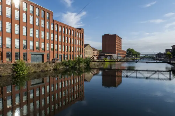 Holyoke, MA-ABD - 24 Ağustos 2025 Sakin bir kanal, kırmızı tuğlalı sanayi binalarını ve Holyoke 'de dağınık mavi bulutların altında bir köprüyü yansıtır.
