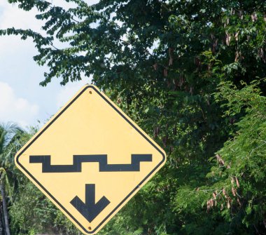 Yellow road sign with graphics and arrow indicating an obstacle designed to decrease speed, in the background tropical trees in Mexico