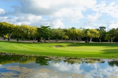 Golf arabasındaki golfçüler, Meksika 'daki tropik iklimde güneşli bir günde gölün yakınındaki son çekim için yeşilin yakınına gidiyorlar.
