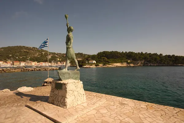Yunanistan 'ın Paxos adasında gün batımı ışığı ve Yunan bayrağıyla birlikte Gaios köyünün marinasının girişindeki Georgios Anemogiannis' in bronz heykeli