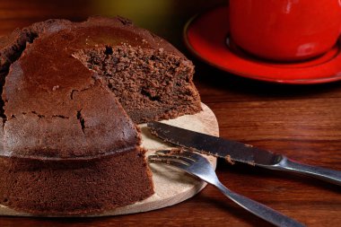 Kesilmiş yuvarlak çikolatalı keke yakın plan. Tahta kesme tahtasının üzerinde yoğurt malzemesi var. Bir bıçak, bir çatal, kırmızı bir fincan ve bir tabak. Koyu ahşap bir masaya yerleştirilmiş..