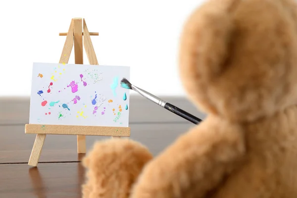 A Teddy bear holding a paint brush is painting colorful spatters on a canvas held by a wooden easel. Dark wooden flooring, white background.