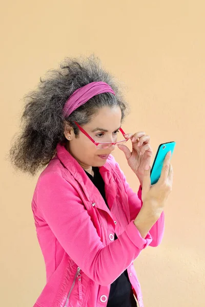 A smiling woman over 50 in pink clothing is checking her mobile phone, A sandy brown wall in the background. Medium shot.