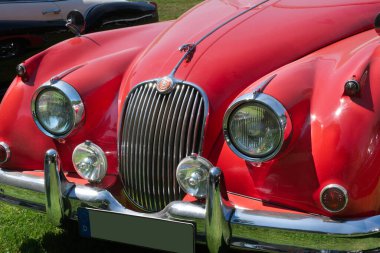 1958 yılından Red Jaguar XK 150 SE OTS otomobili. Ön detaylar Jaguar maskotunu, logoyu, ızgarayı, araba ışıklarını ve tamponu gösteriyor. Açık hava, güneş ışığı.