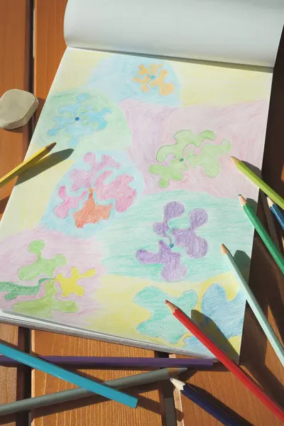 A colorful floral naive pencil drawing with abstract flowers on a sketch book. Colored pencils scattered around the book. Items placed on a table under sunlight.
