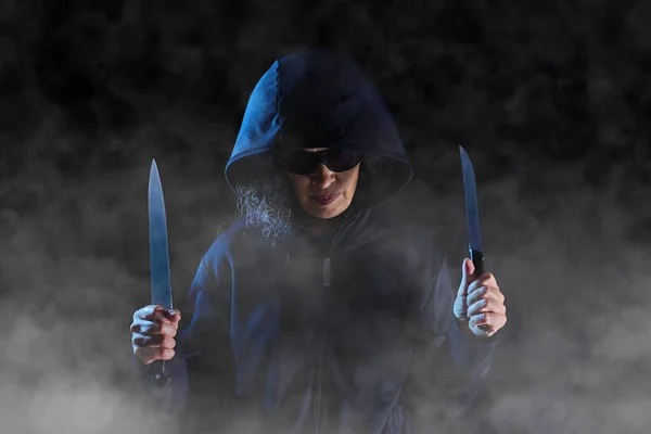 A person wearing a dark hood and dark sunglasses is holding a knife in each hand. Person surrounded by fog. Dark background.