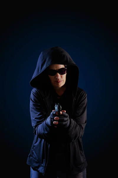 A person wearing a dark hood and dark sunglasses is pointing a gun to the viewer. Dark cyan-blue gradient background.