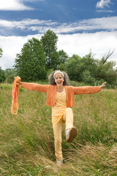 Laughing woman over 50 making a big movement outdoor in the nature. She has graying hair and wears orange clothing. Trees and sky in the background.