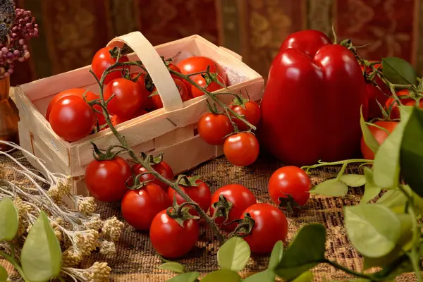Sepette kırmızı kiraz domatesleri, kırmızı dolma biber, kurutulmuş bitkiler ve antika görünümlü masa örtüsüne yerleştirilmiş yeşil bir bitki..