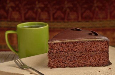 Tahta kesme tahtasının üzerinde bir dilim çikolatalı pasta. Arkadaki yeşil kupa. Ögelerin arkasında koyu bir retro arkaplan.