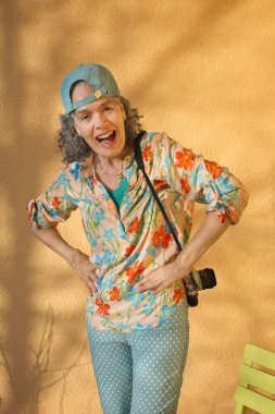 Yaz kıyafetleri içindeki mutlu Boomer kadın poz veriyor. Omzunun bir tarafında retro kamerası var. Şeftali gibi turuncu bir arka planda gün ışığıyla oynadım..