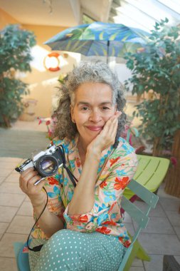 Kıvırcık saçlı, 61 yaşındaki Boomer kadın kış bahçesinde gülümsüyor. Bir elinde klasik bir film kamerası tutuyor..