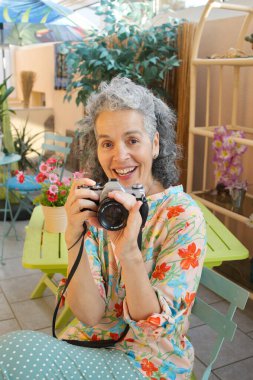 Altmış bir yaşında kıvırcık saçlı bir kadın kış bahçesinde oturmuş gülümsüyor. Bir elinde eski bir film kamerası tutuyor..