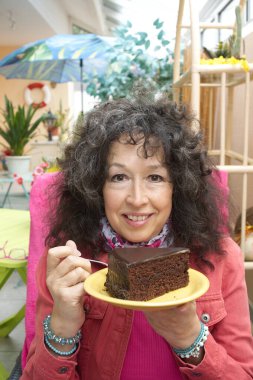 60 yaşını geçmiş bir kadın elinde çikolatalı pastayla mutlu mesut oturuyor. Gri kıvırcık saçlı. Arka planda kış bahçesi.