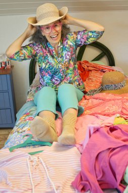 Hasır şapka takan gülen kadın renkli yaz kıyafetleriyle dolu bir yatakta oturuyor..