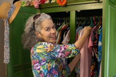 Altmış bir yaşındaki gülümseyen kadın yeşil gardırobunda kıyafet seçiyor. Gri kıvırcık saçları var ve renkli bir gömlek giyiyor..