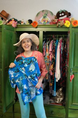 Gülümseyen kadın yeşil gardırobundan mavi Hawaii bluzu seçiyor. Hasır şapka ve yaz kıyafetleri giyiyor..