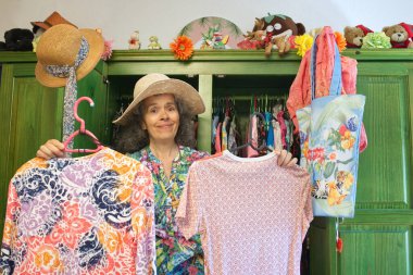 Dolabının önünde asılı iki gömlek tutan olgun bir kadın var. Yüzünde kararsız bir ifade.