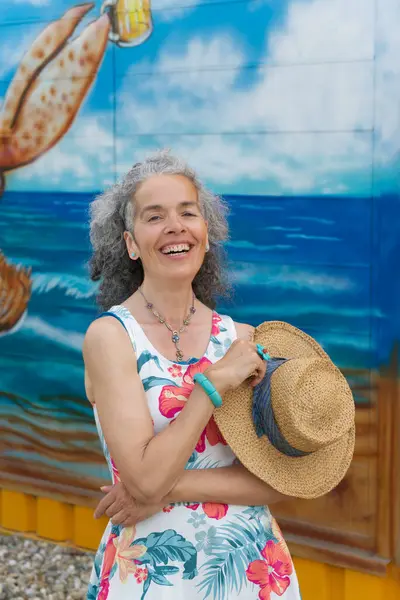 Kıvırcık saçlı, gülen olgun bir kadın yaz elbisesi giyiyor ve elinde hasır bir şapka tutuyor. Renkli arkaplan.