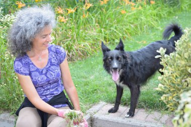 Bahçesinde çalışırken köpeğine gülümseyen olgun bir kadın. Arka planda zambaklar.