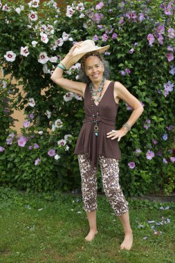 Bahçede poz veren gülümseyen olgun kadın. Başının üzerinde hasır bir şapka tutuyor ve hayvan desenli giysiler ve turkuaz mücevherler takıyor. Arka planda çiçek açan bir Hibiscus Syriacus.