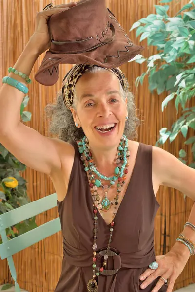 Kafasına şapka geçirmiş olgun bir kadına gülüyordu. Gri kıvırcık saçları var ve kahverengi bir yaz kıyafeti giyiyor. Arkaplanda bitkiler ve bir sazlık ekran.