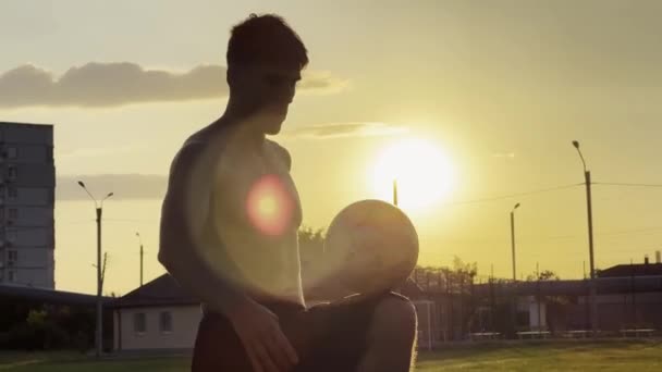 日没のスタジアムでサッカーボールをジャグリングする若者 グリーンフィールドでボールを蹴るプロサッカー選手 スポーツマンは バックグラウンドで日光でメドウでトリックを実践しています フリースタイルフットボール — ストック動画