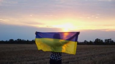 Arpa çayırında yürüyüp Ukrayna bayrağını dalgalandıran mutlu bir kadın. Arka planda güzel bir günbatımında mavi-sarı pankartıyla Ukraynalı bir kadın. Savaşın sonu..