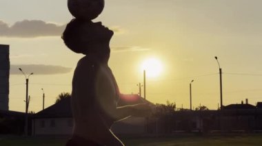 Genç adam gün batımında stadyumda futbol topuyla hokkabazlık yapıyor. Yeşil sahada profesyonel futbolcu topa vuruyor. Sporcu arka planda güneş ışığıyla çayırda numaralar yapıyor. Serbest stil futbol