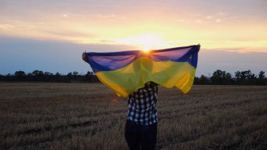 Arpa çayırında yürüyüp Ukrayna bayrağını dalgalandıran mutlu bir kadın. Arka planda güzel bir günbatımında mavi-sarı pankartıyla Ukraynalı bir kadın. Savaşın sonu.
