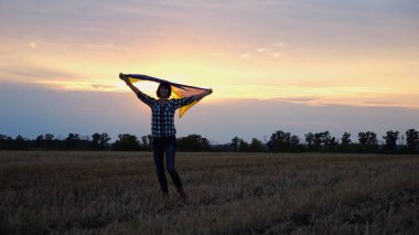 Arpa çayırında yürüyüp Ukrayna bayrağını dalgalandıran mutlu bir kadın. Arka planda güzel bir günbatımında mavi-sarı pankartıyla Ukraynalı bir kadın. Savaşın sonu.
