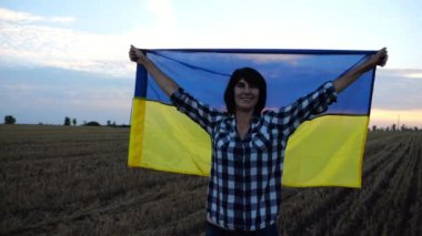 Mutlu kadın gün batımında mavi-sarı bir pankartıyla buğday tarlasında duran kameraya bakıyor. Ukraynalı gülümseyen kadın Ukrayna bayrağının üzerinde gündoğumunda arpa çayırında poz veriyor.