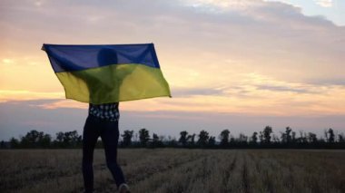 Ukraynalı bir kadın, kafasının üstünde Ukrayna bayrağıyla buğday tarlasında güzel günbatımının arka planına doğru gidiyor. Rus saldırısına karşı zafer sembolü olarak ulusal bayrak taşıyan kadın