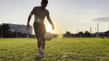 Genç adam gün batımında stadyumda futbol topuyla hokkabazlık yapıyor. Yeşil sahada profesyonel futbolcu topa vuruyor. Sporcu arka planda güneş ışığıyla çayırda numaralar yapıyor. Serbest stil futbol