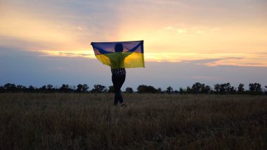 Ukraynalı bir kadın, kafasının üstünde Ukrayna bayrağıyla buğday tarlasında güzel günbatımının arka planına doğru gidiyor. Rus saldırısına karşı zafer sembolü olarak ulusal bayrak taşıyan kadın
