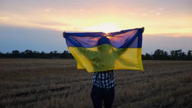 Arpa çayırında yürüyüp Ukrayna bayrağını dalgalandıran mutlu bir kadın. Arka planda güzel bir günbatımında mavi-sarı pankartıyla Ukraynalı bir kadın. Savaşın sonu.