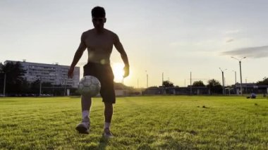 Genç adam gün batımında stadyumda futbol topuyla hokkabazlık yapıyor. Yeşil sahada profesyonel futbolcu topa vuruyor. Sporcu arka planda güneş ışığıyla çayırda numaralar yapıyor. Serbest stil futbol