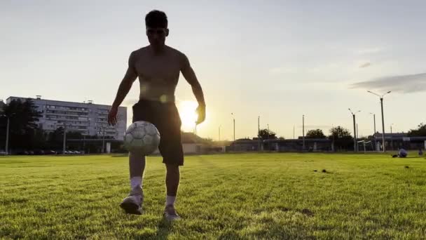 年轻男子日落时在体育场抛接足球 职业足球运动员在绿地踢球 运动员在草地上练习杂技 背景是阳光 自由式足球 — 图库视频影像