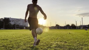 Genç adam gün batımında stadyumda futbol topuyla hokkabazlık yapıyor. Yeşil sahada profesyonel futbolcu topa vuruyor. Sporcu arka planda güneş ışığıyla çayırda numaralar yapıyor. Serbest stil futbol