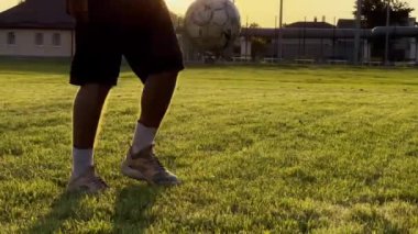 Genç adam gün batımında stadyumda futbol topuyla hokkabazlık yapıyor. Yeşil sahada profesyonel futbolcu topa vuruyor. Sporcu arka planda güneş ışığıyla çayırda numaralar yapıyor. Serbest stil futbol