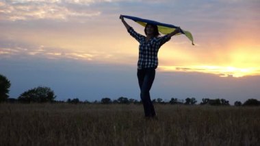 Arpa çayırında yürüyüp Ukrayna bayrağını dalgalandıran mutlu bir kadın. Arka planda güzel bir günbatımında mavi-sarı pankartıyla Ukraynalı bir kadın. Savaş konsepti bitti.