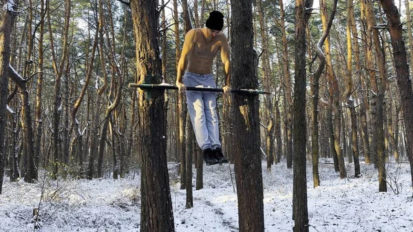 Strong and muscular man doing exercises on horizontal bar at winter forest. Young sportsman muscling up at beautiful nature. Athletic guy training outdoor. Concept of sport and active lifestyle.